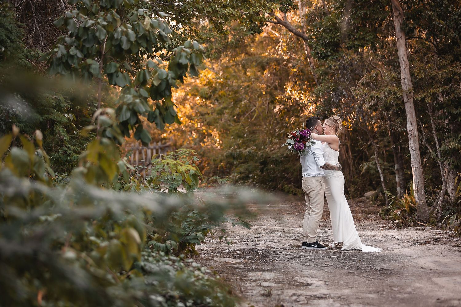 Bride and groom embracing on a rustic dirt path surrounded by lush autumn foliage in a romantic outdoor wedding photo.