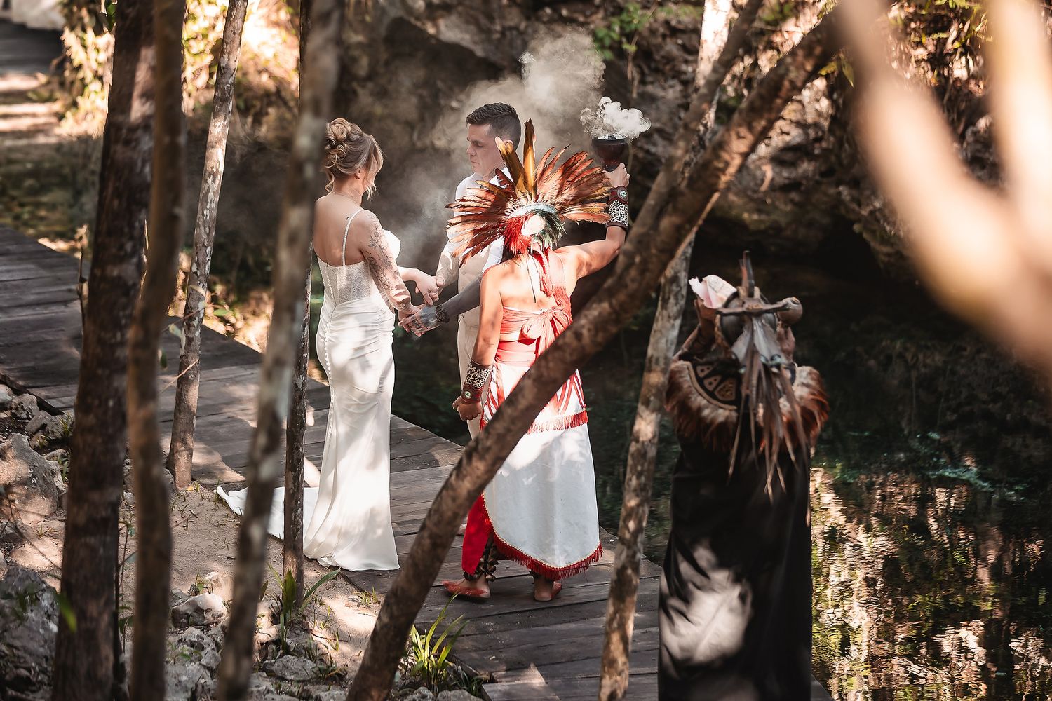 Couple in traditional attire exchange vows with officiant in a lush forest setting during an outdoor wedding ceremony.