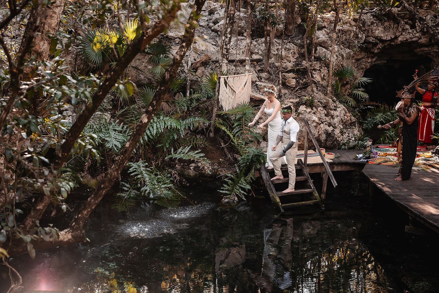 Bride and groom crossing a wooden bridge over a lush jungle cenote in Mexico.