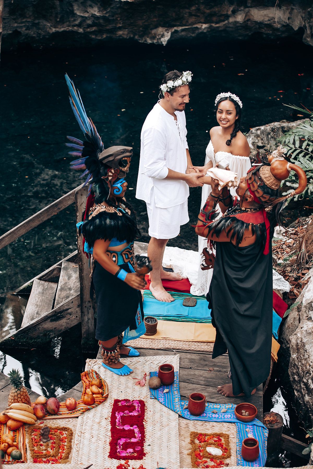 Wedding ceremony with traditional performers and ceremonial items arranged on colorful textiles by a dark pool.