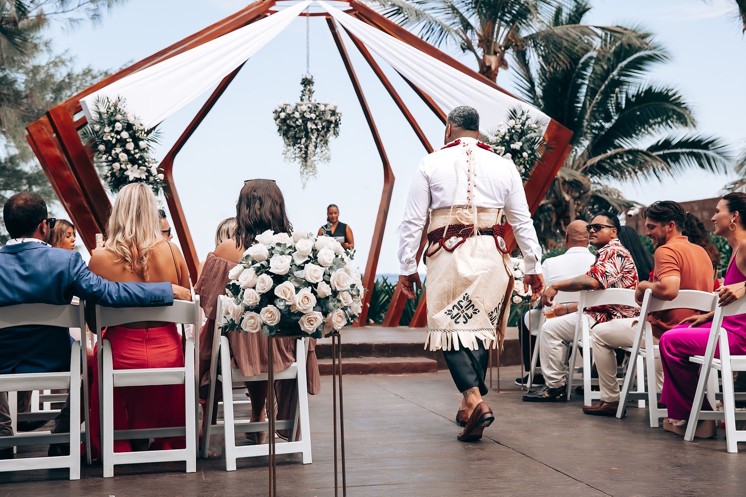 Outdoor wedding ceremony setup with wooden arch and white chairs under palm trees.