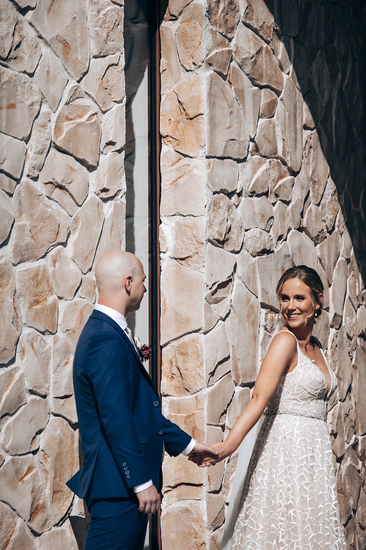 A couple stands in front of a stone wall, the bride wearing a lace wedding dress.