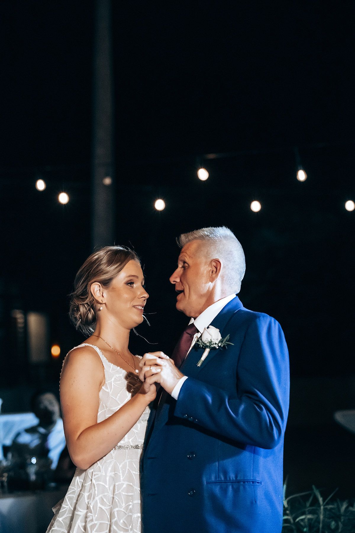 A bride and groom dance together under a night sky with twinkling lights.