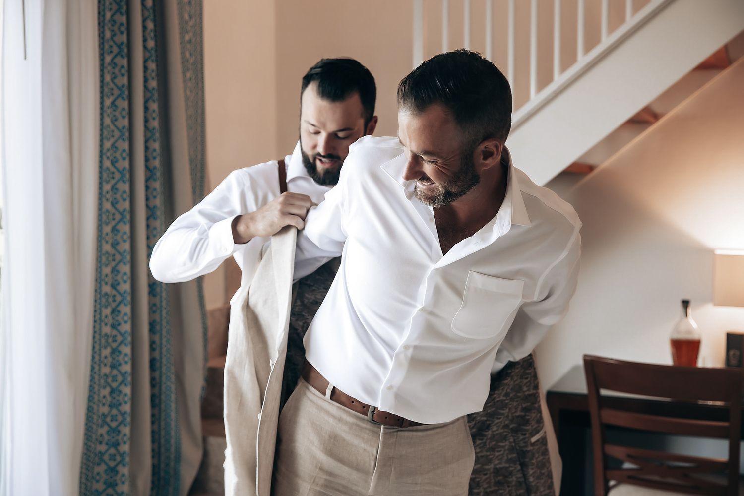 Two men in white shirts are helping each other get dressed for a wedding.