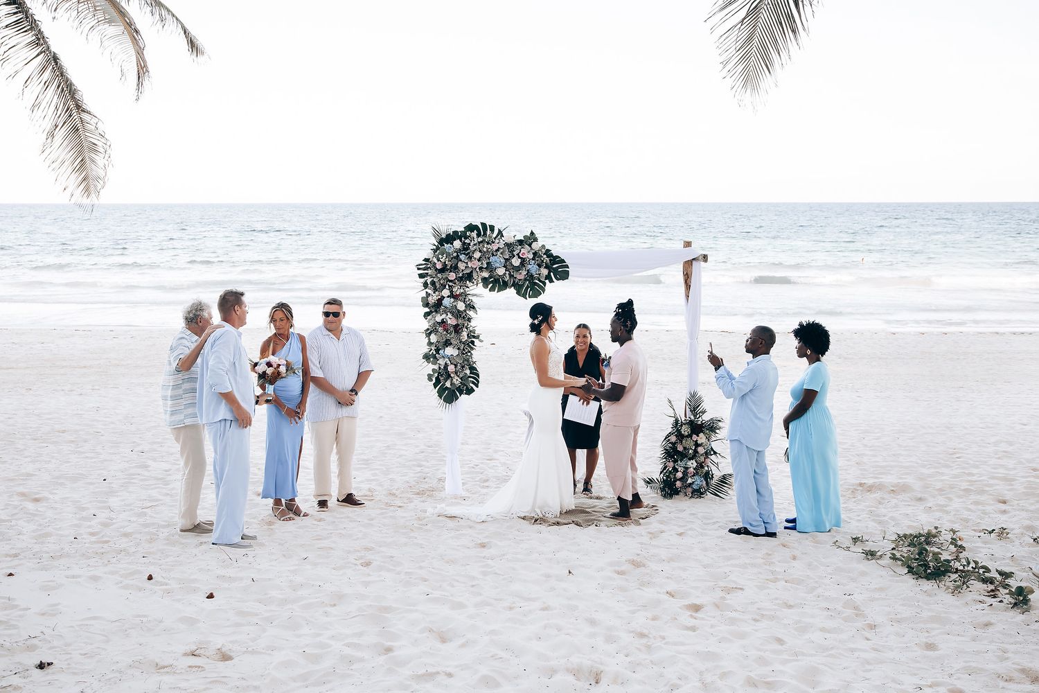 A wedding ceremony takes place on a beach, with the couple and their wedding party standing in front of a floral arch.