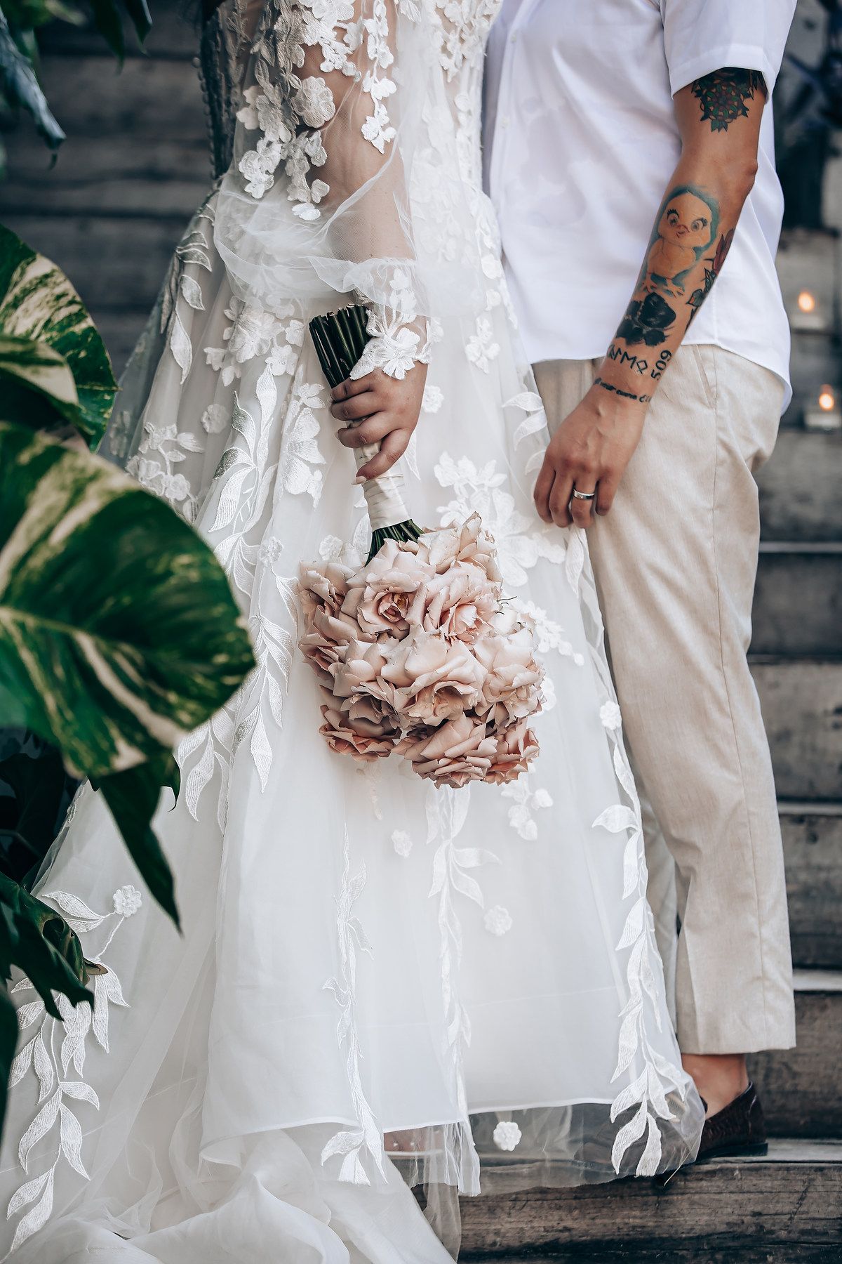 Close-up details of a bride's lace wedding dress and bouquet with blush pink flowers on stone steps.