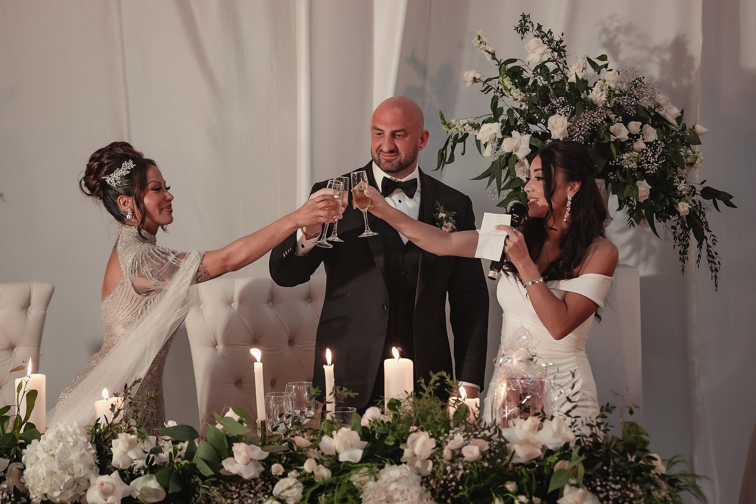 Wedding guests enjoying a reception dinner with laughter and toasts at an elegant white-decorated venue.