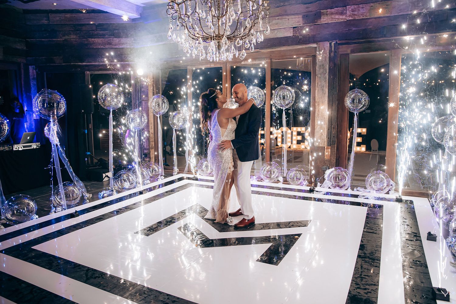 A couple shares their first dance on an illuminated black and white checkered dance floor under crystal chandeliers.