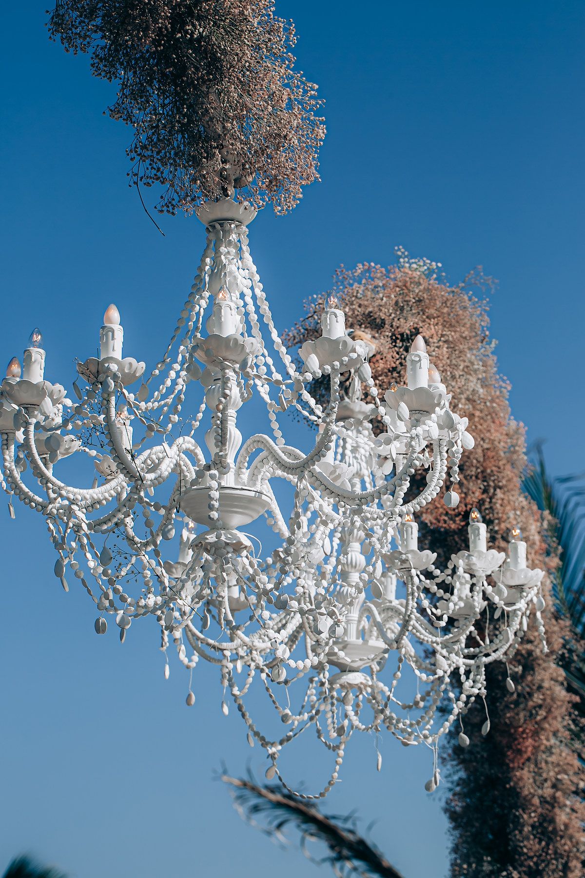 Ornate white crystal chandelier with cascading glass beads hangs against bright blue sky with tree branches.