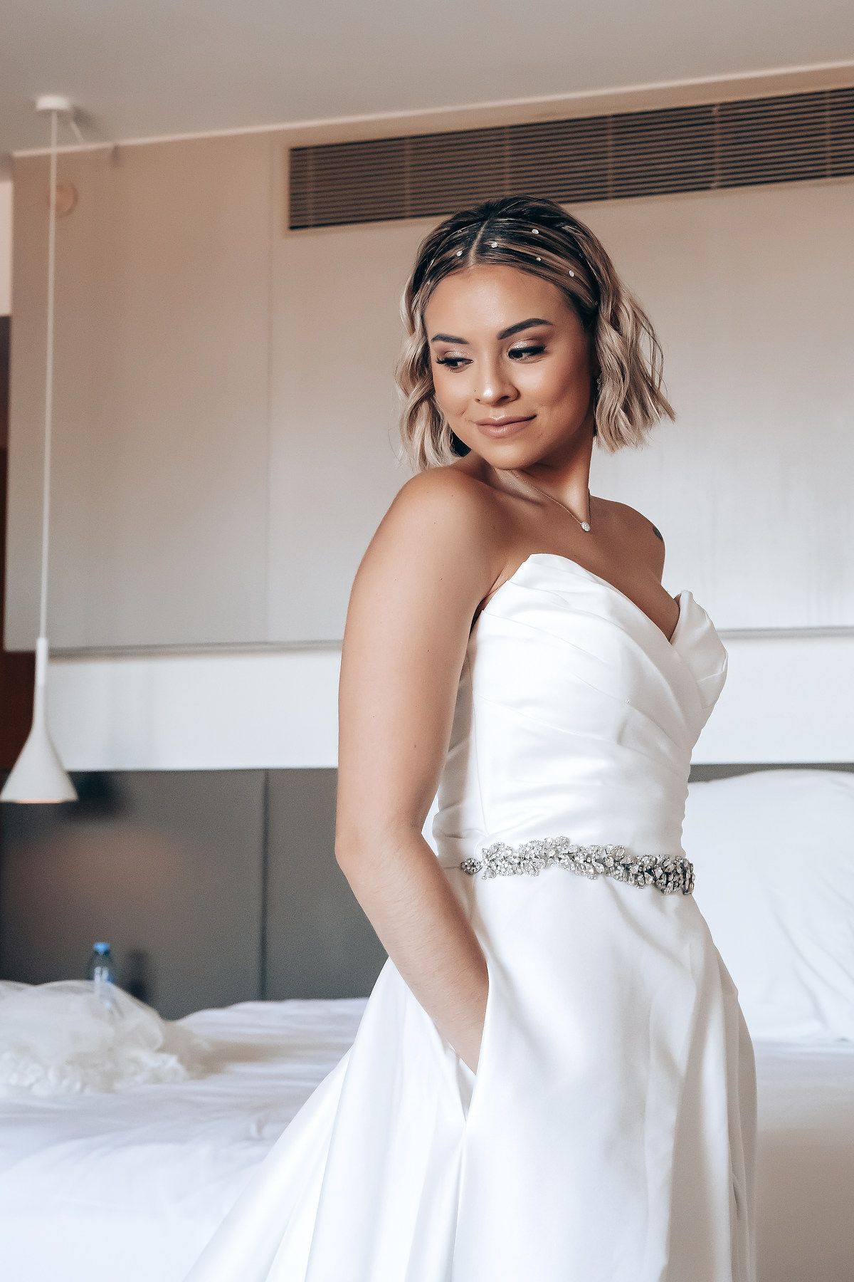 Bride in elegant white wedding gown with crystal belt and short wavy hair poses in hotel room.