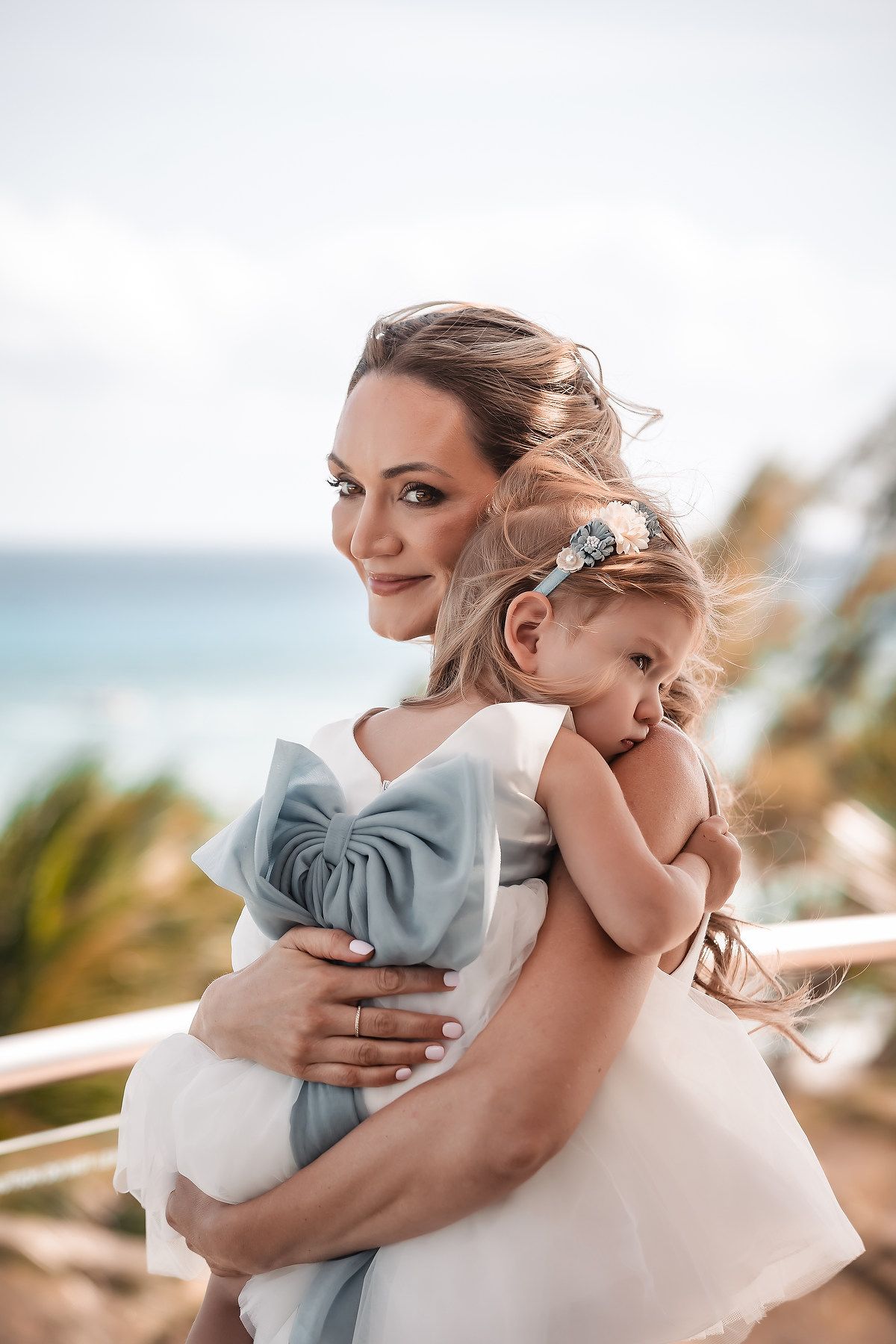 A tender embrace between parent and child with windswept hair against a tropical beach backdrop.