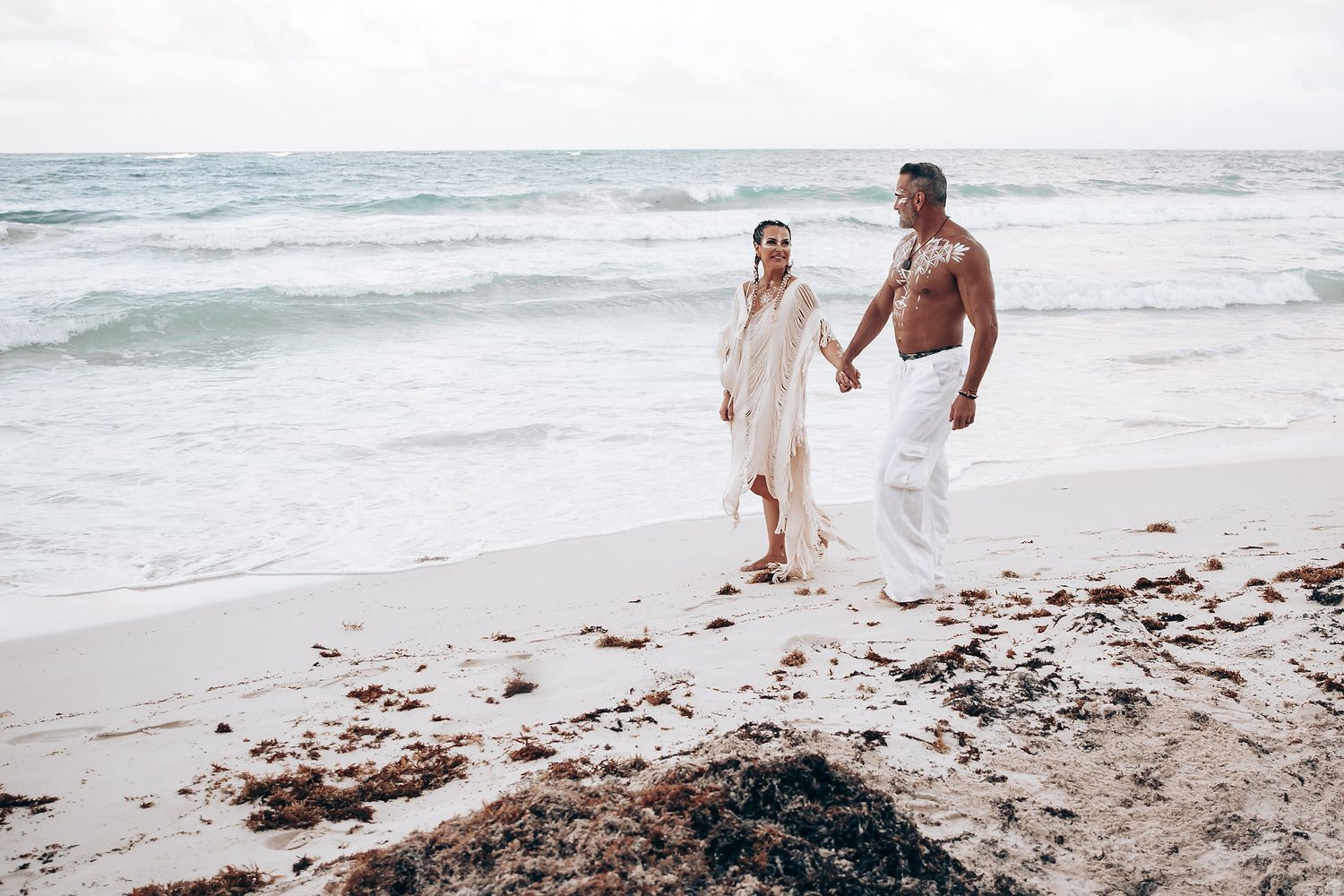 A couple walking along the shoreline wearing white beach attire.