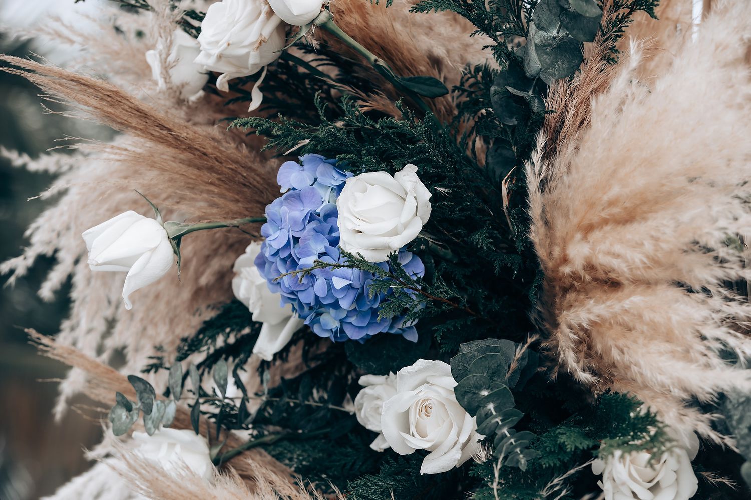 A wedding floral arrangement featuring blue hydrangeas, white roses and pampas grass against a dark background.