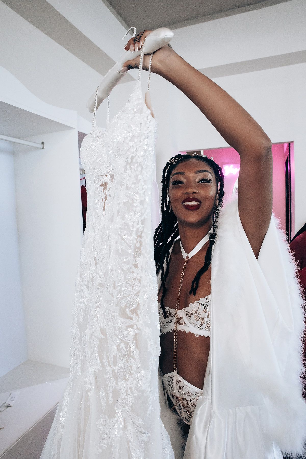 A joyful person wearing a white lace wedding dress poses playfully in a bright indoor space.