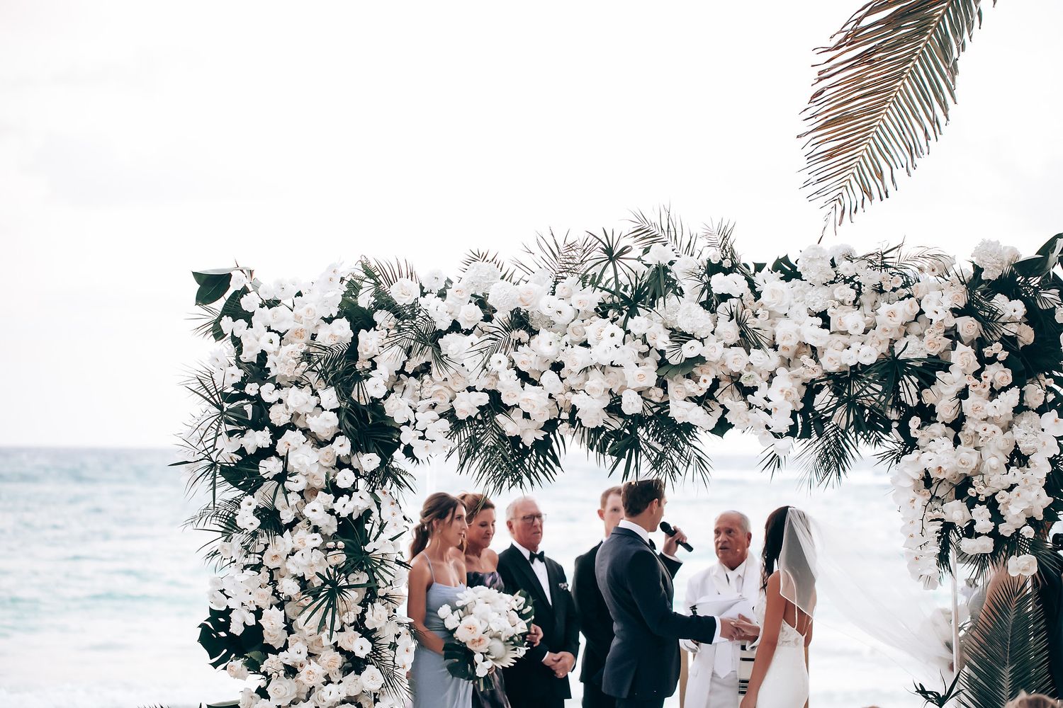 Beach wedding ceremony with elaborate white flower arch and ocean backdrop.