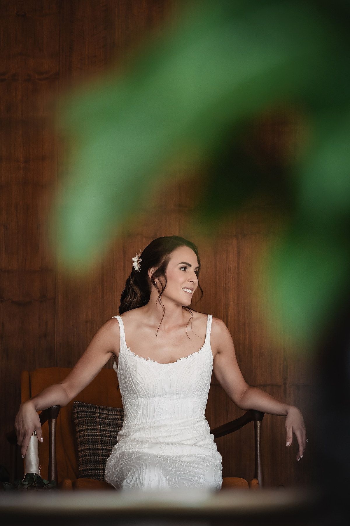 A person in a white wedding dress poses dramatically against a wooden wall with green foliage in the background.