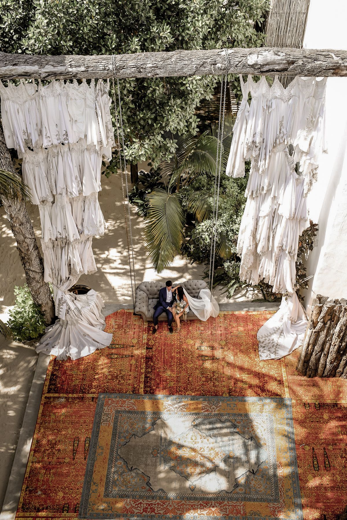 Collection of white wedding dresses hanging outdoors on display with vintage rugs underneath.