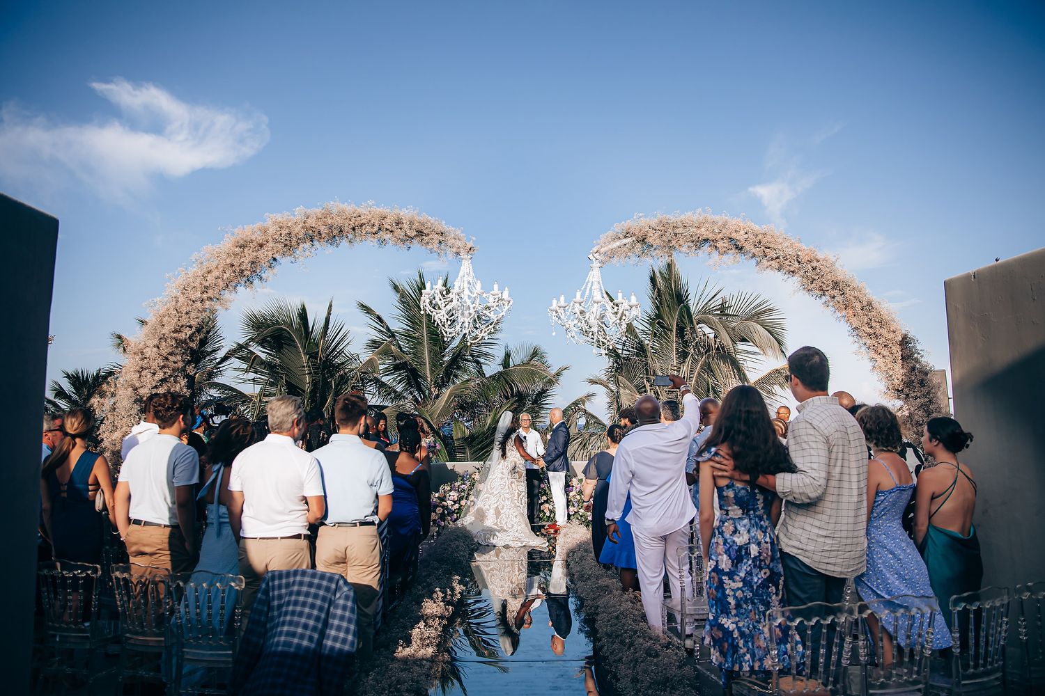 Beach wedding ceremony setup with floral arches and white chairs against a blue sky.