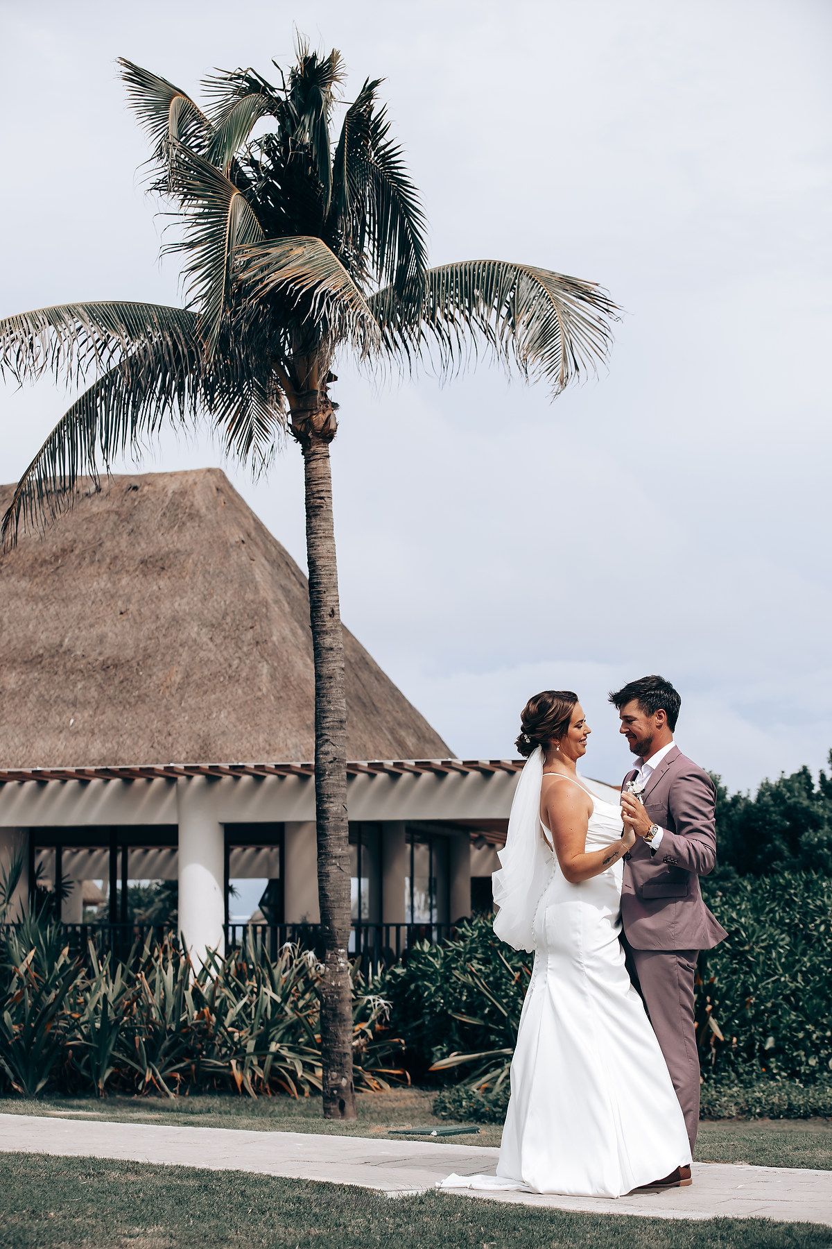 Wedding couple poses together against a tropical backdrop with thatched-roof bungalows and palm trees.