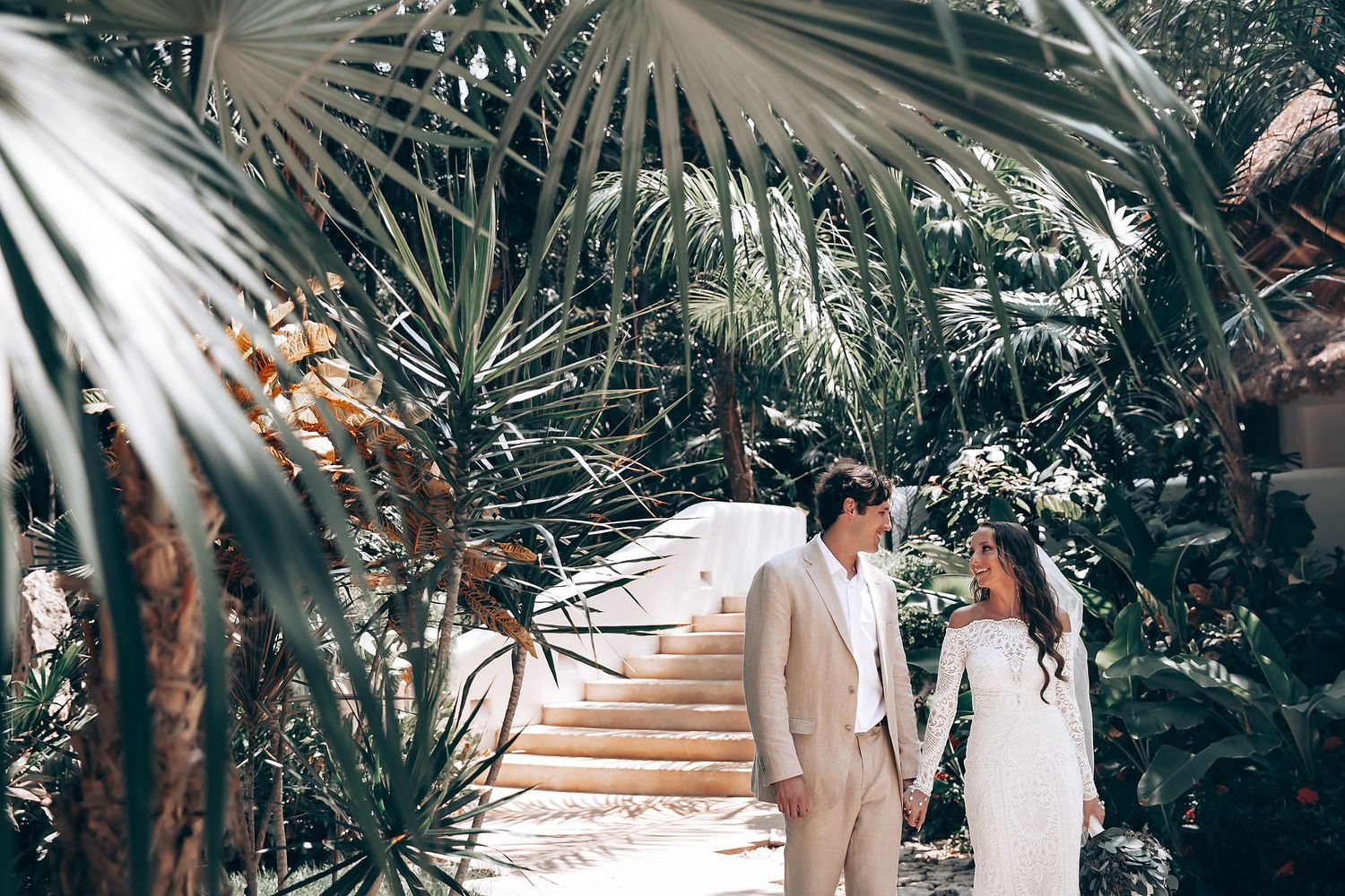 Newlyweds ascending white staircase surrounded by tropical foliage and palm fronds.