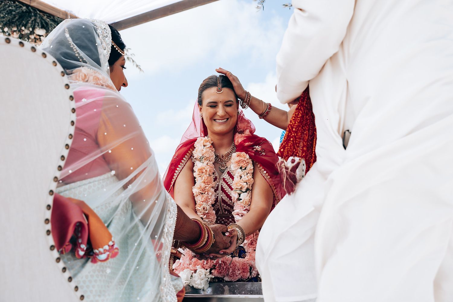 Wedding ceremony participants gather under a white canopy with floral decorations by the ocean.
