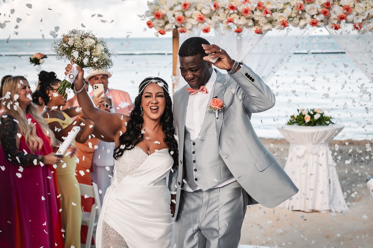 Joyful celebration at beach wedding with flower petals being tossed in the air.