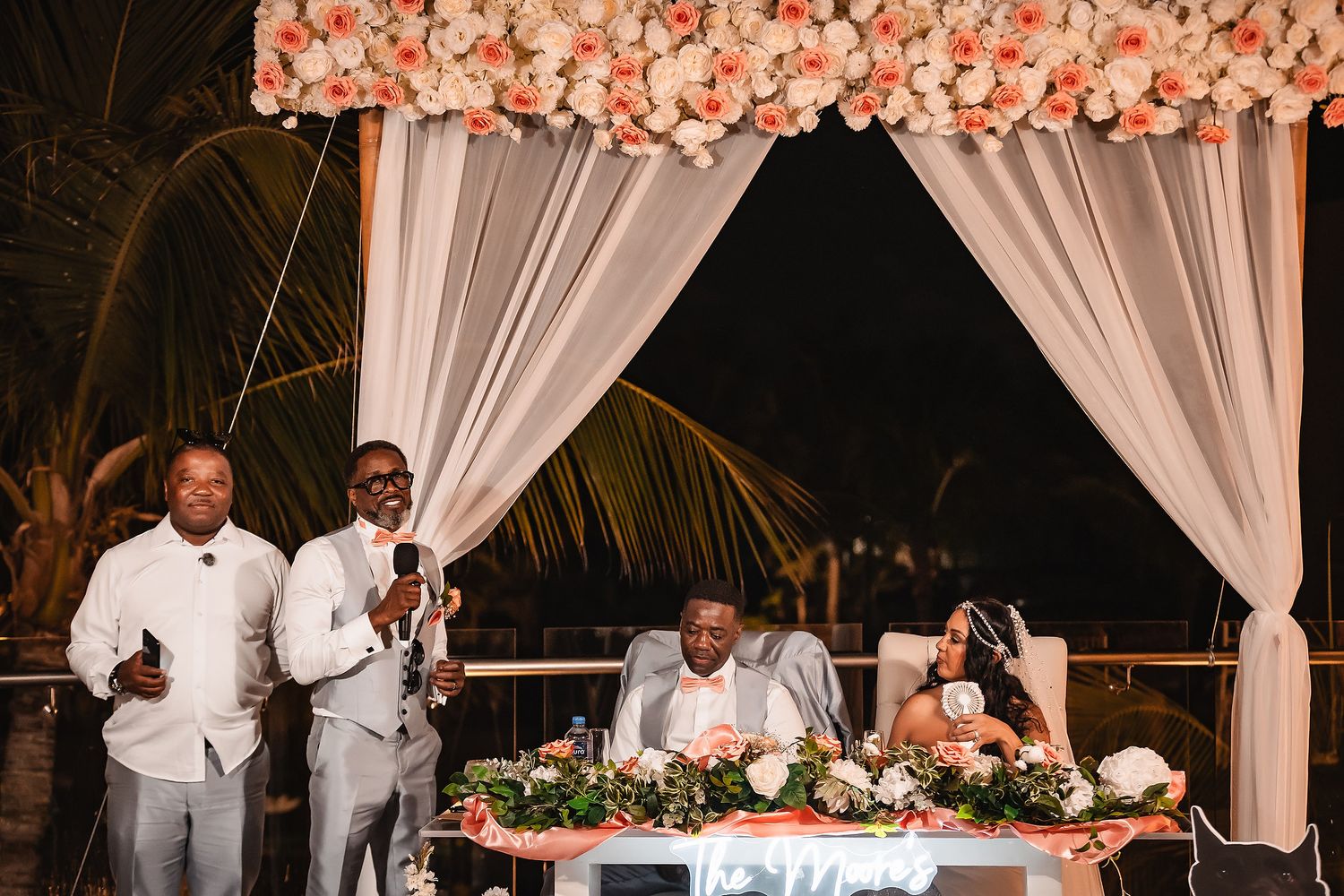 Wedding reception table decorated with coral flowers and white draping under evening lighting.