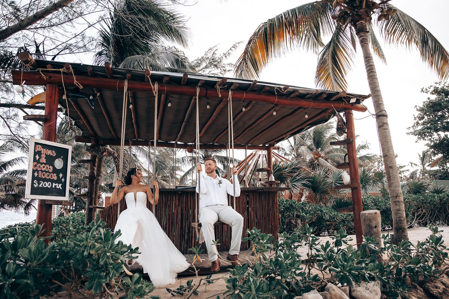 A couple in wedding attire poses playfully on a rustic wooden platform surrounded by tropical landscaping.