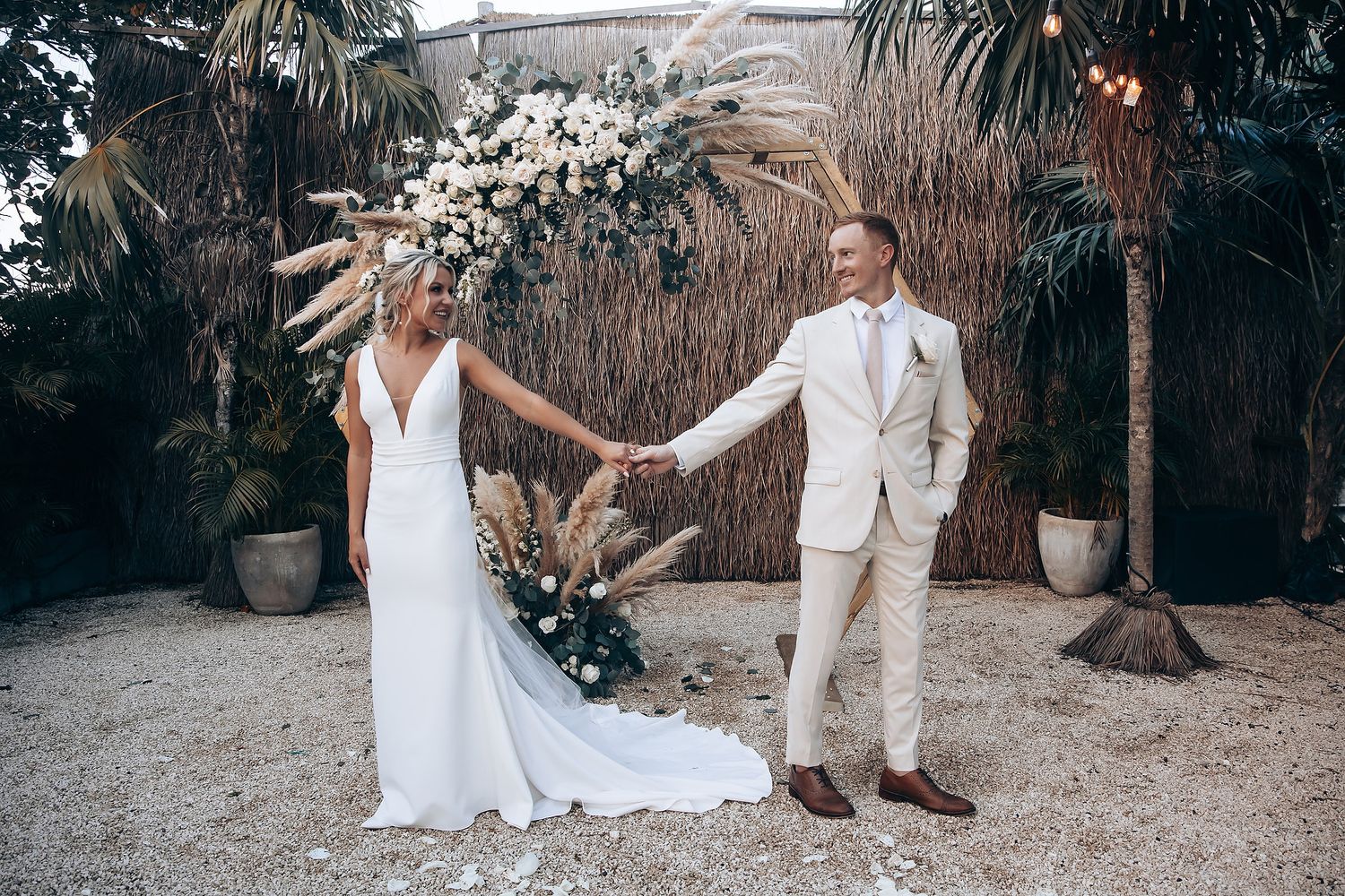 A couple in wedding attire holding hands under tropical palm trees and white floral arrangements.