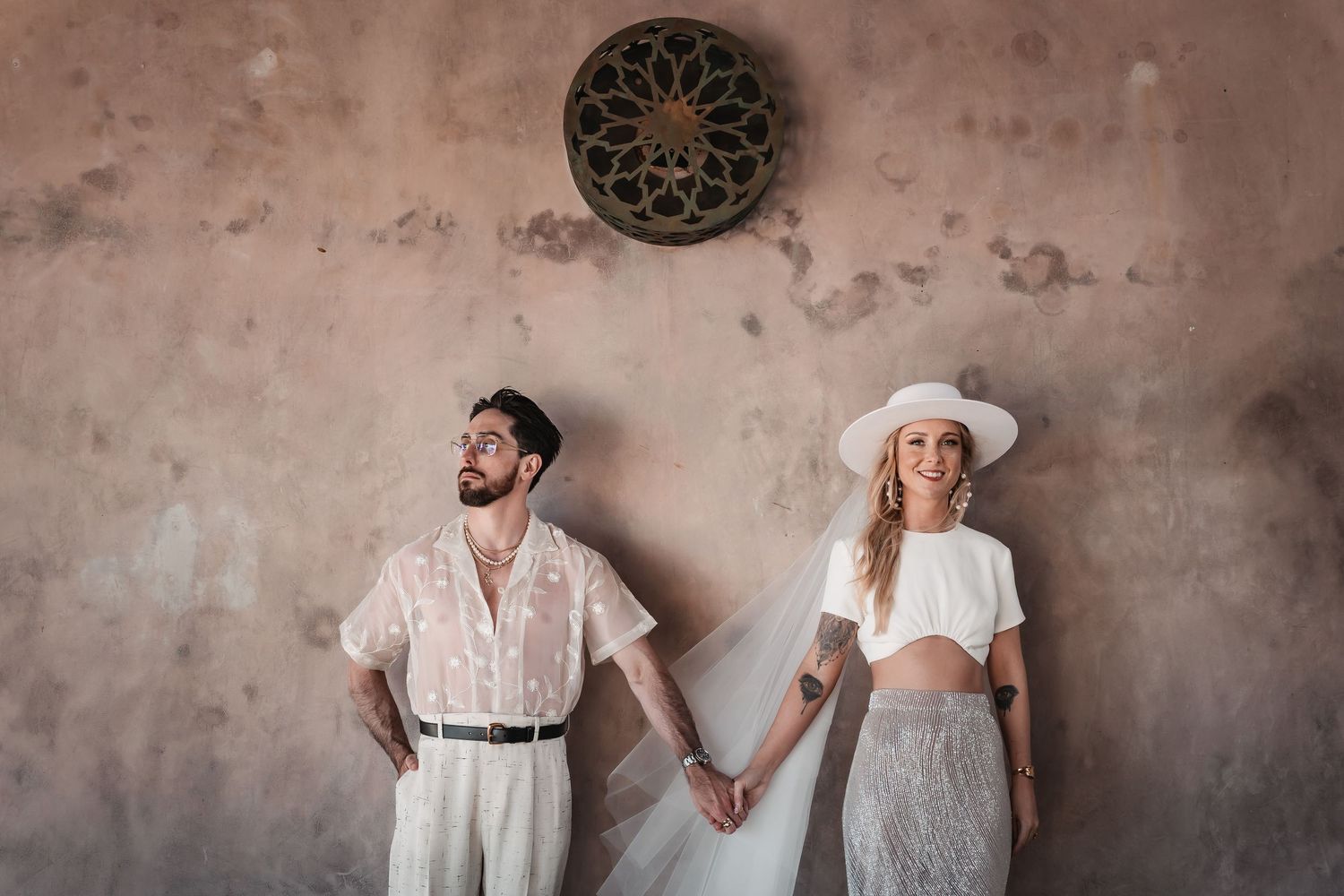 Stylish couple in white outfits poses against textured wall with ornate decor.