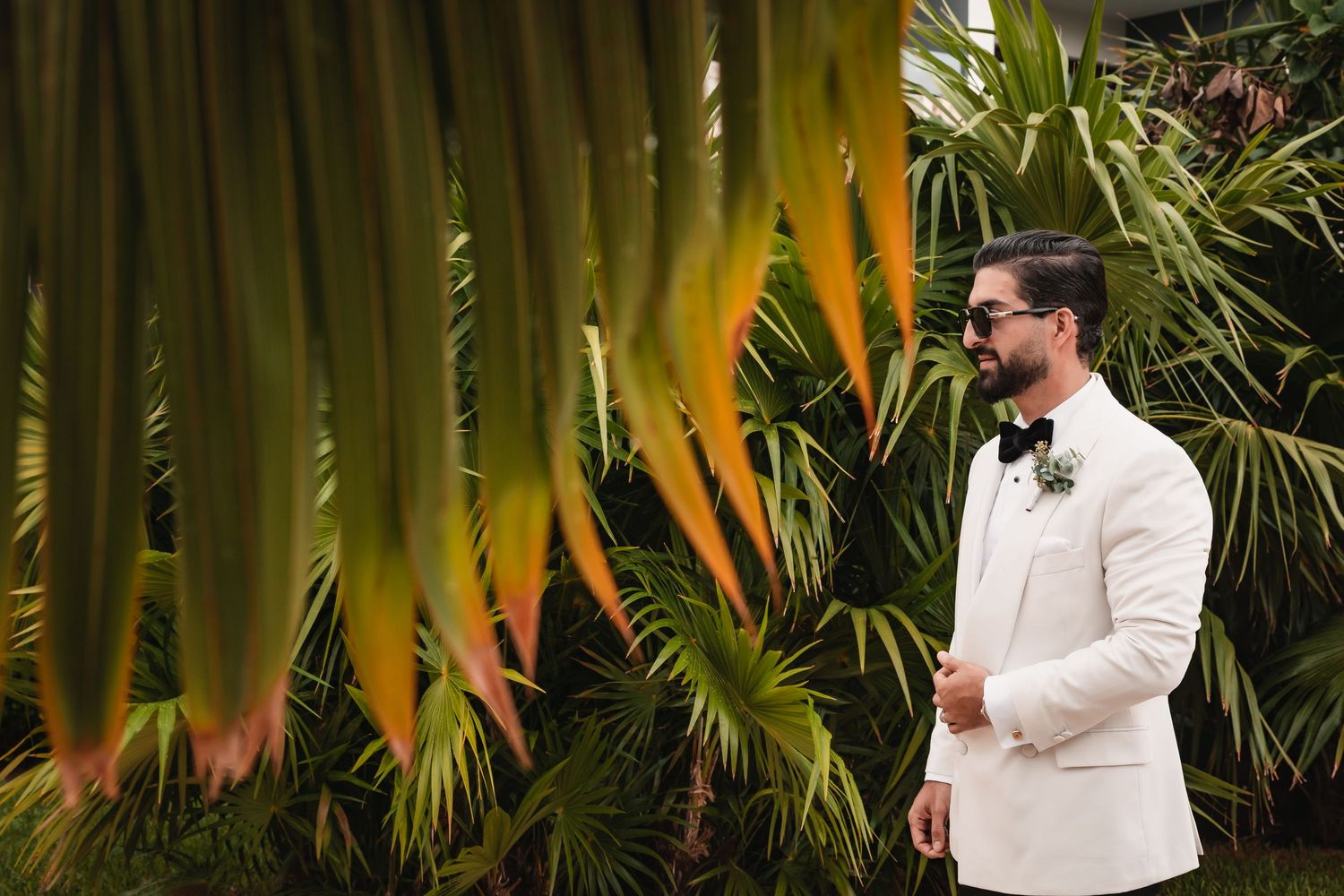 Wedding party members in white suits pose against tropical palm fronds in a garden setting.
