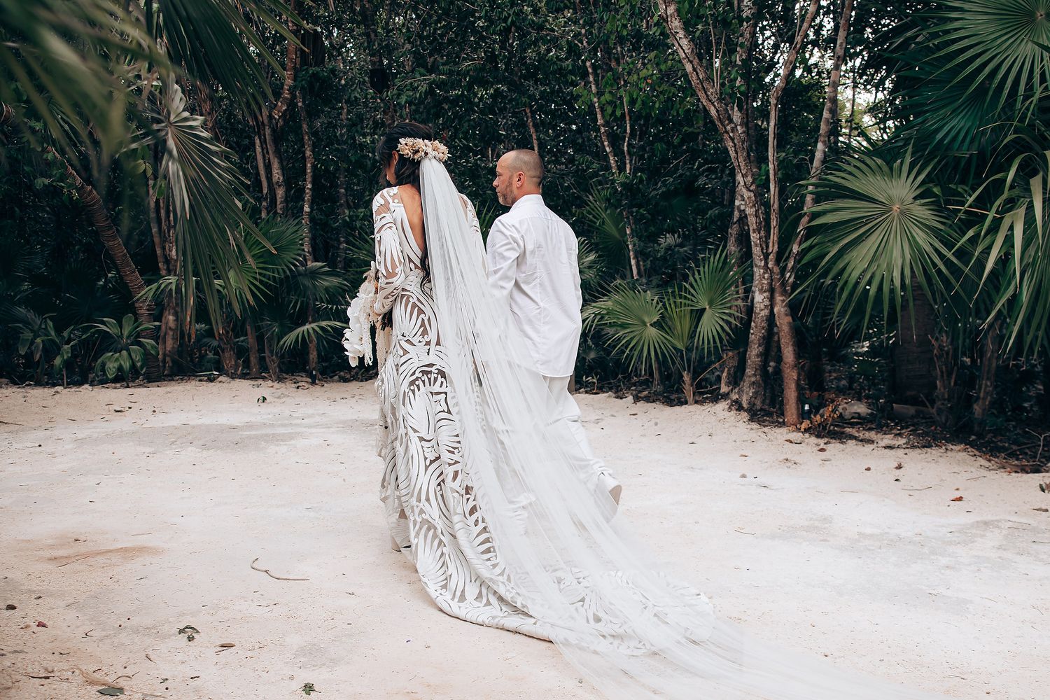 A couple in wedding attire walks hand in hand through a lush tropical garden path with palm trees.