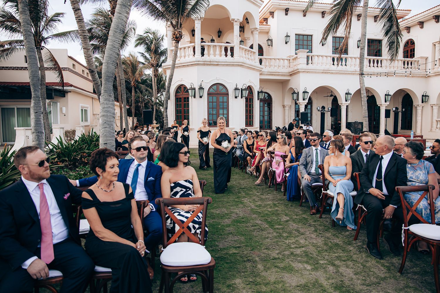 Guests gather on a lawn for an outdoor wedding ceremony at an elegant Mediterranean-style mansion with arches and balconies.