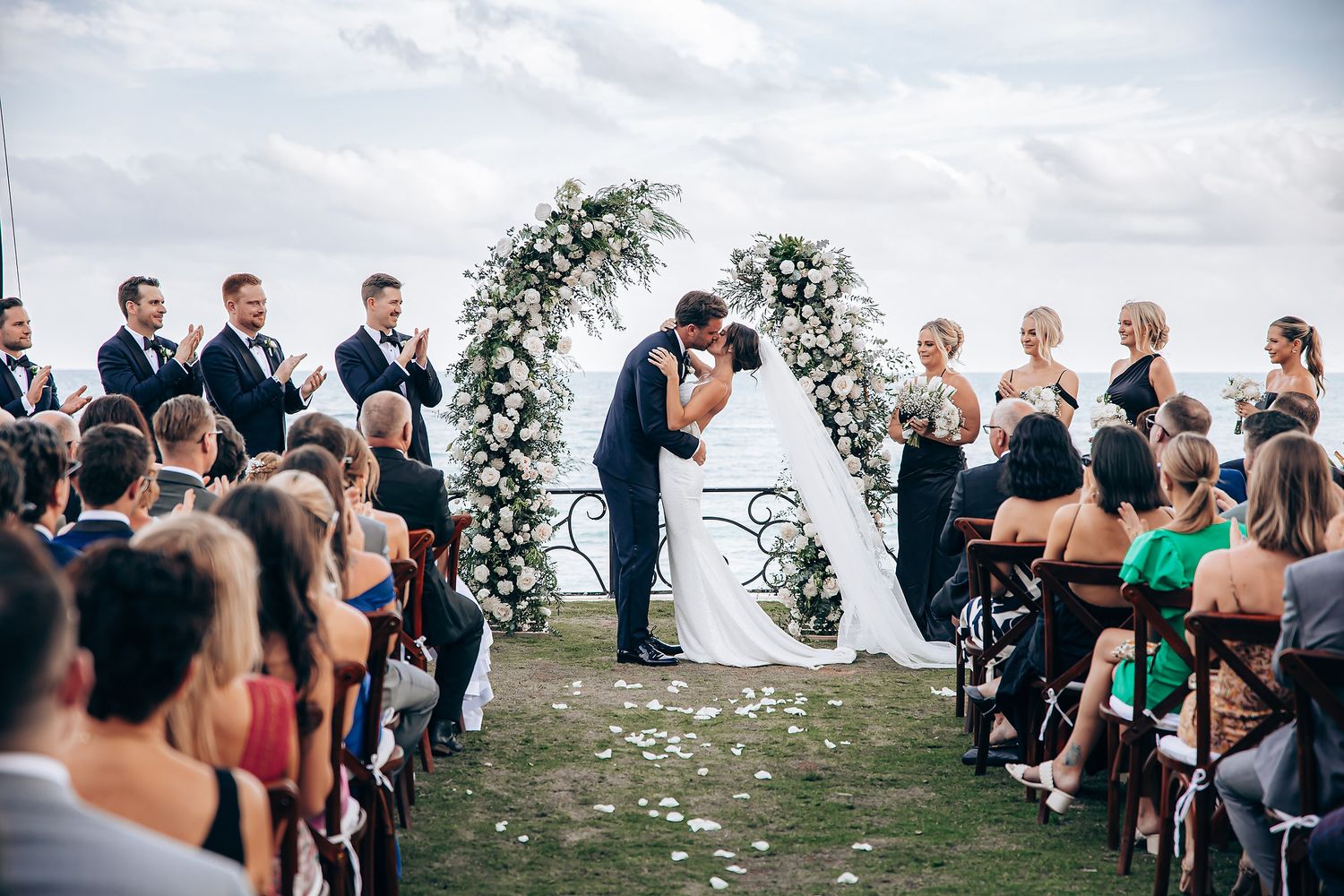Wedding guests gathered outdoors for a ceremony with palm trees and ocean views in the background.