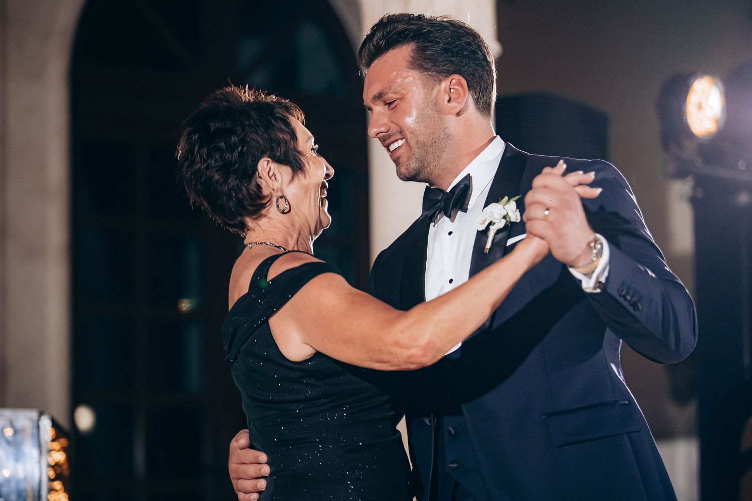 Couple dancing together at a wedding reception in formal attire with romantic lighting in the background.