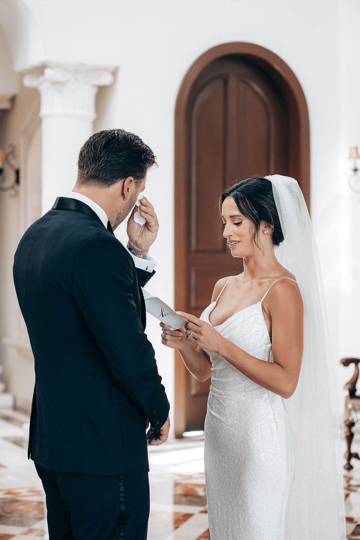 An emotional wedding ceremony moment as a bride in white lace dress and groom in black suit exchange vows indoors.