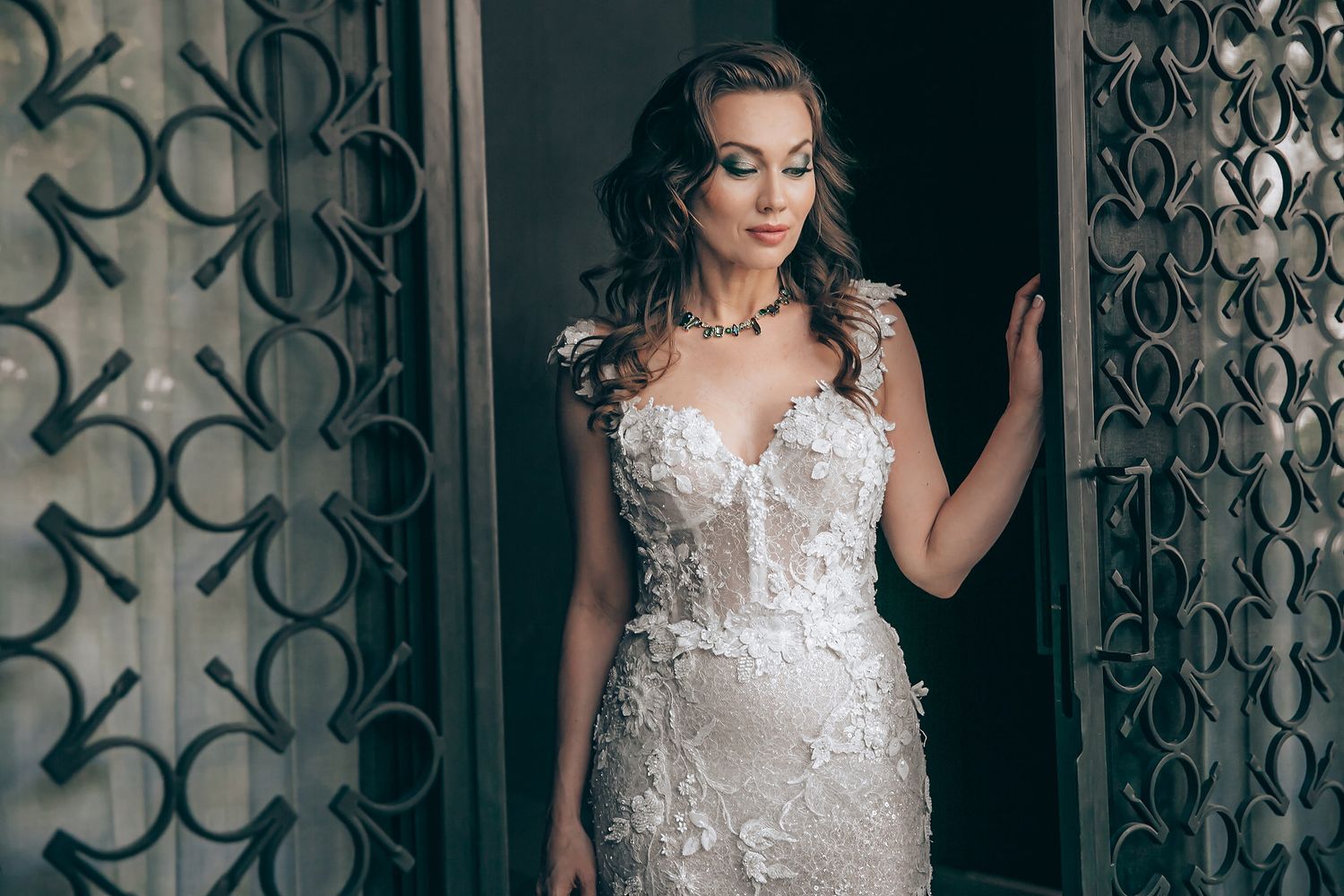 A bride in an elegant white lace wedding dress poses against an ornate dark patterned door.