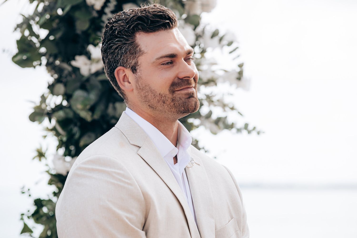 Groom in beige suit jacket smiles during outdoor wedding ceremony against white backdrop.