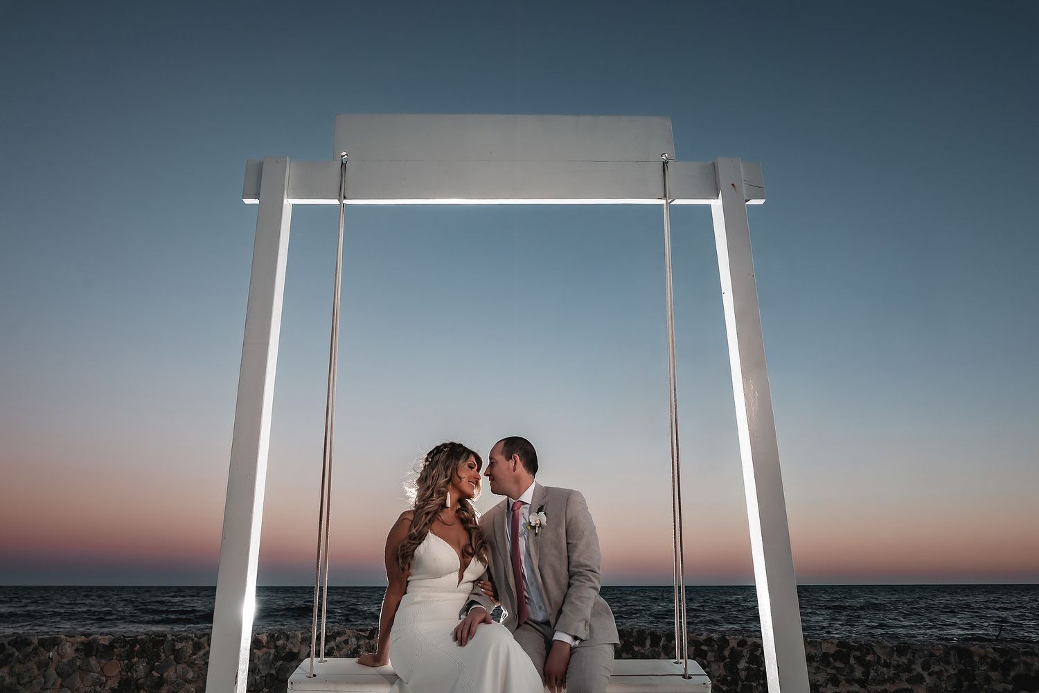 Bride and groom share a romantic moment on a swing at sunset by the ocean during their beach wedding ceremony.