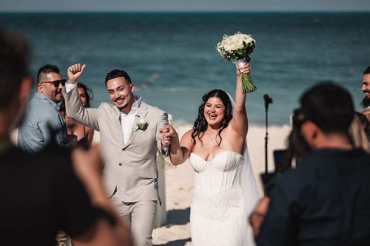 Joyful newlyweds celebrate on a beach wedding, bride raising bouquet, groom holding bottle, guests cheering behind them.