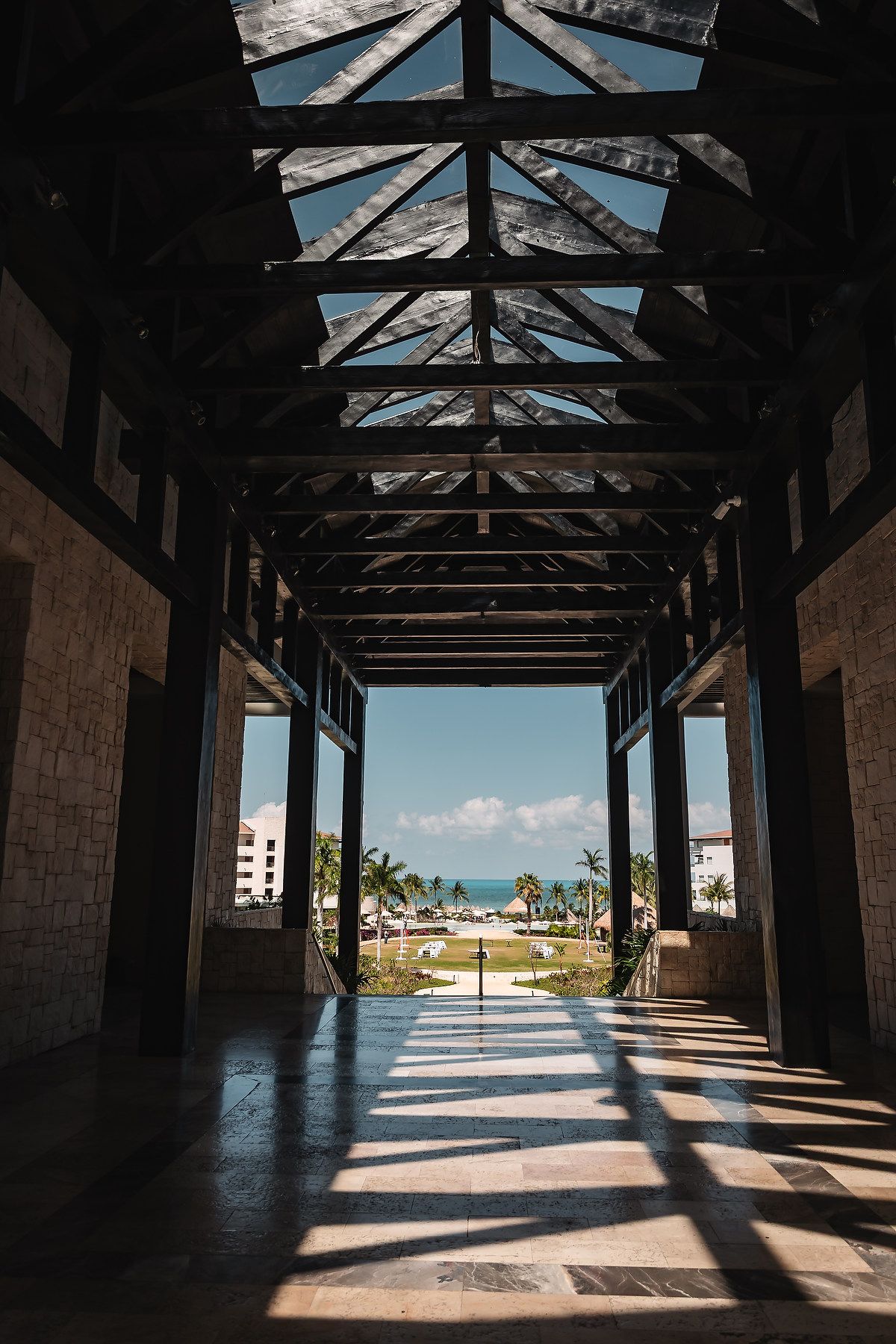 Sunlit covered walkway with wooden beam ceiling leading to tropical resort grounds with palm trees and ocean view.