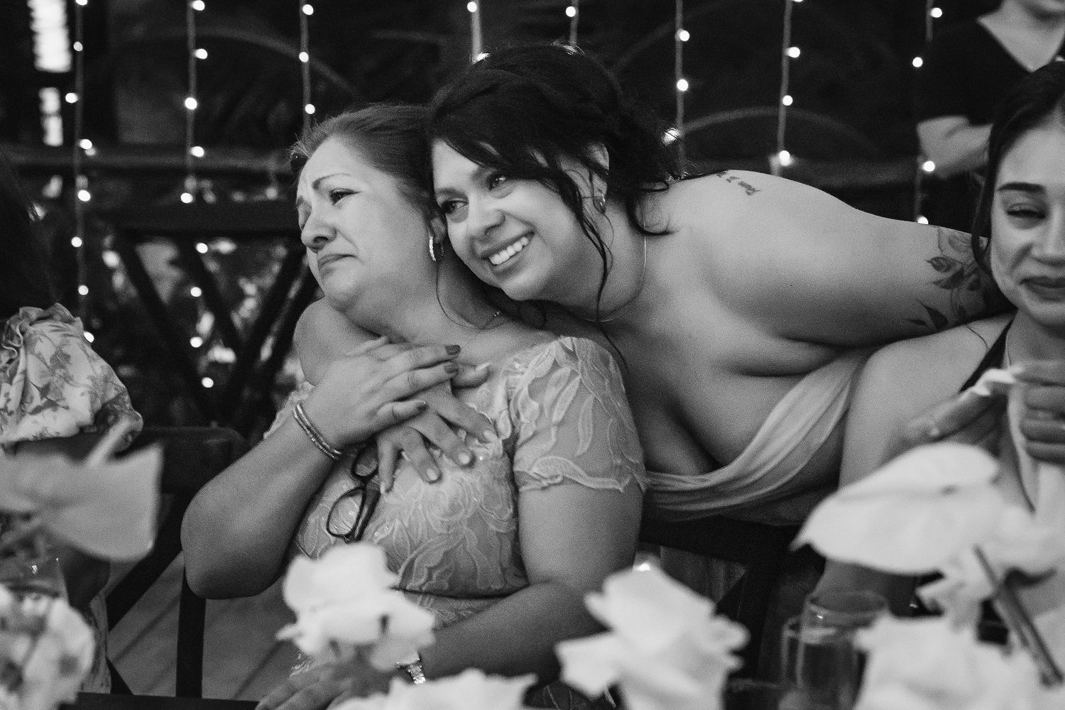 Black and white photo of two women hugging and smiling at a wedding reception with fairy lights in background.