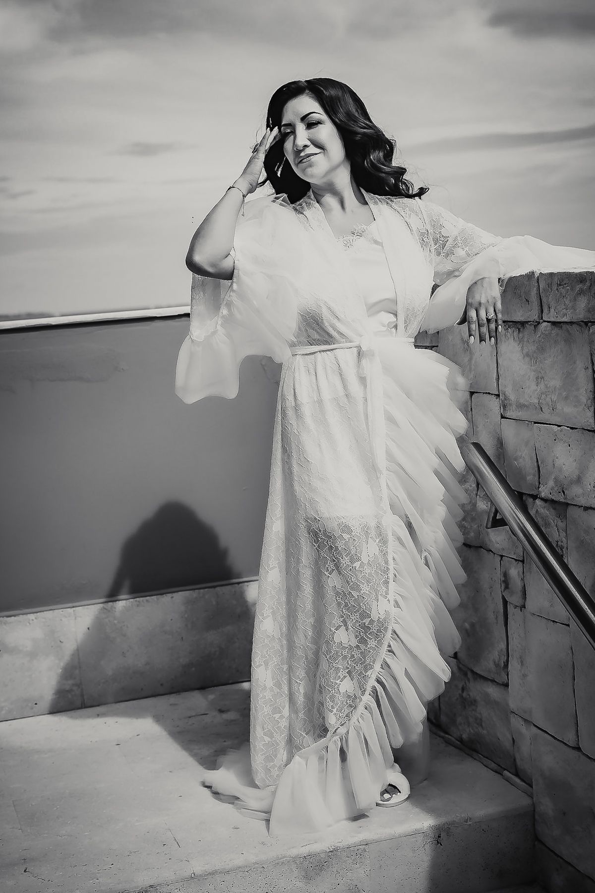 Black and white photo of a woman in an elegant ruffled white gown posing on stone steps outdoors.