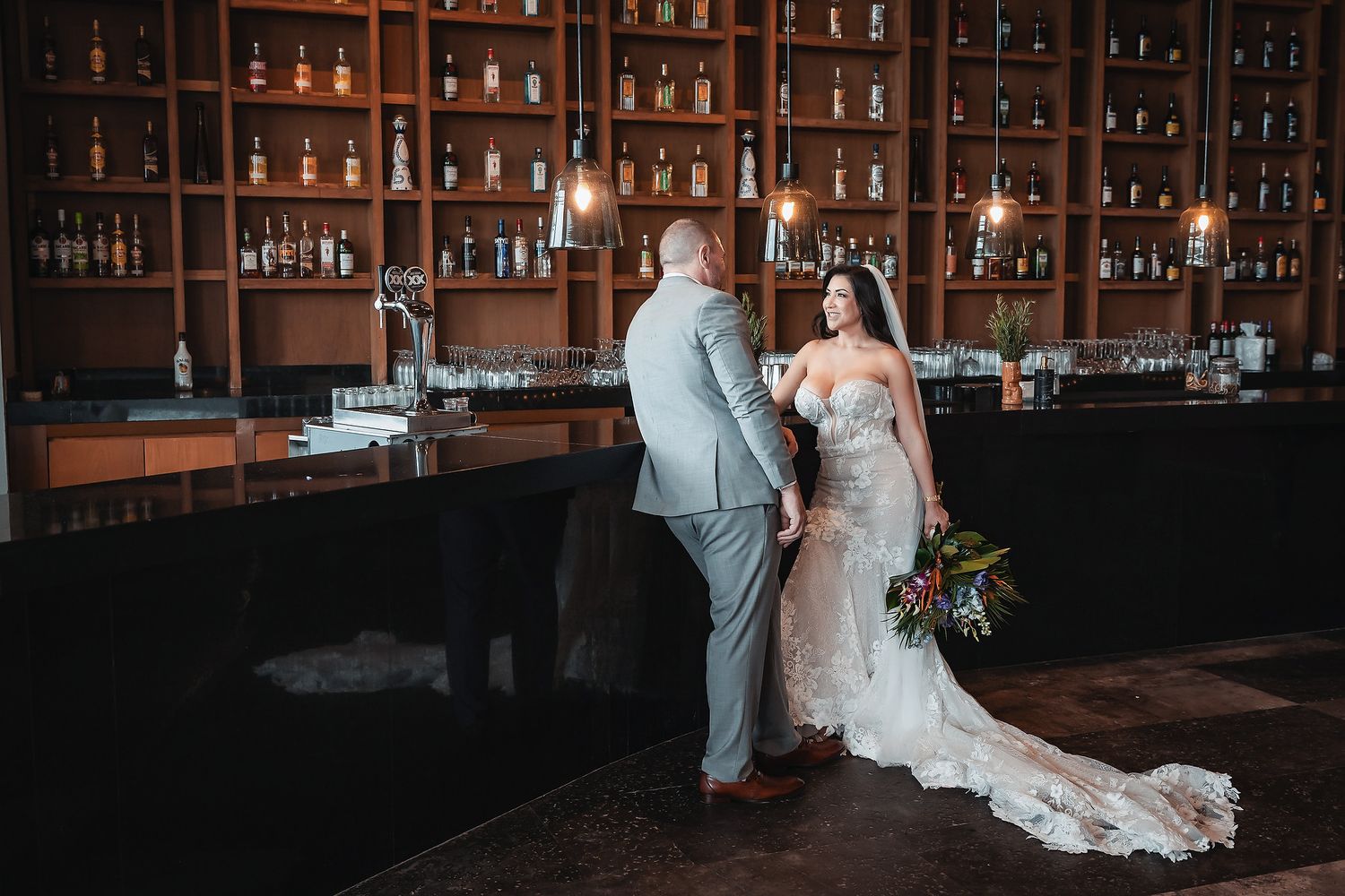Bride and groom share a romantic moment at an elegant bar with wood shelving and bottle displays on their wedding day.