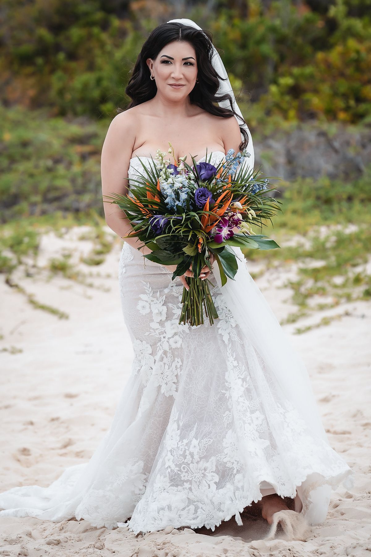 Bride in strapless lace mermaid gown holding tropical bouquet with purple and orange flowers on sandy beach.