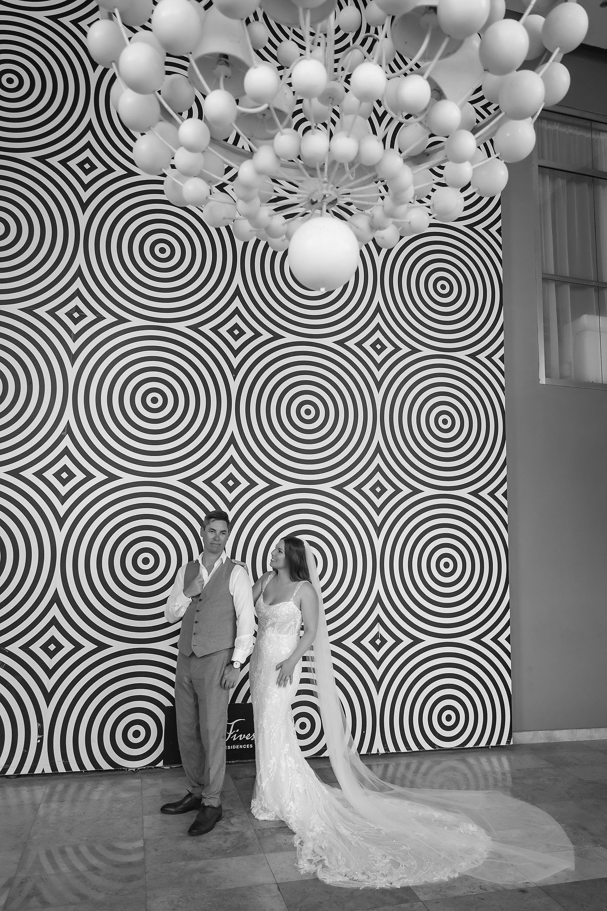 Black and white wedding photo of bride and groom posing before bold geometric circle wall with elegant balloon chandelier.