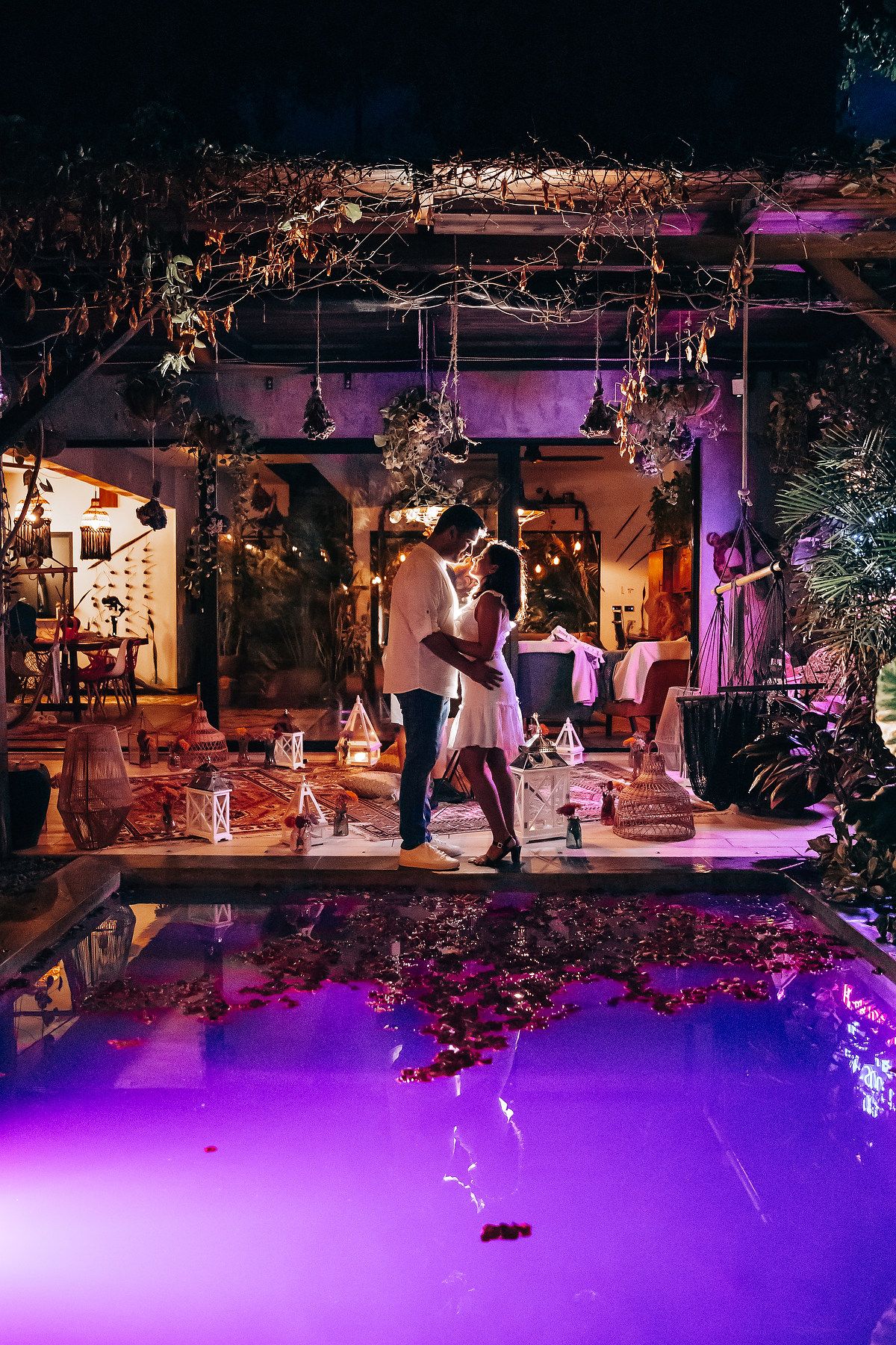 Romantic couple embracing poolside at night under a pergola with purple lighting and rose petals floating in the water.
