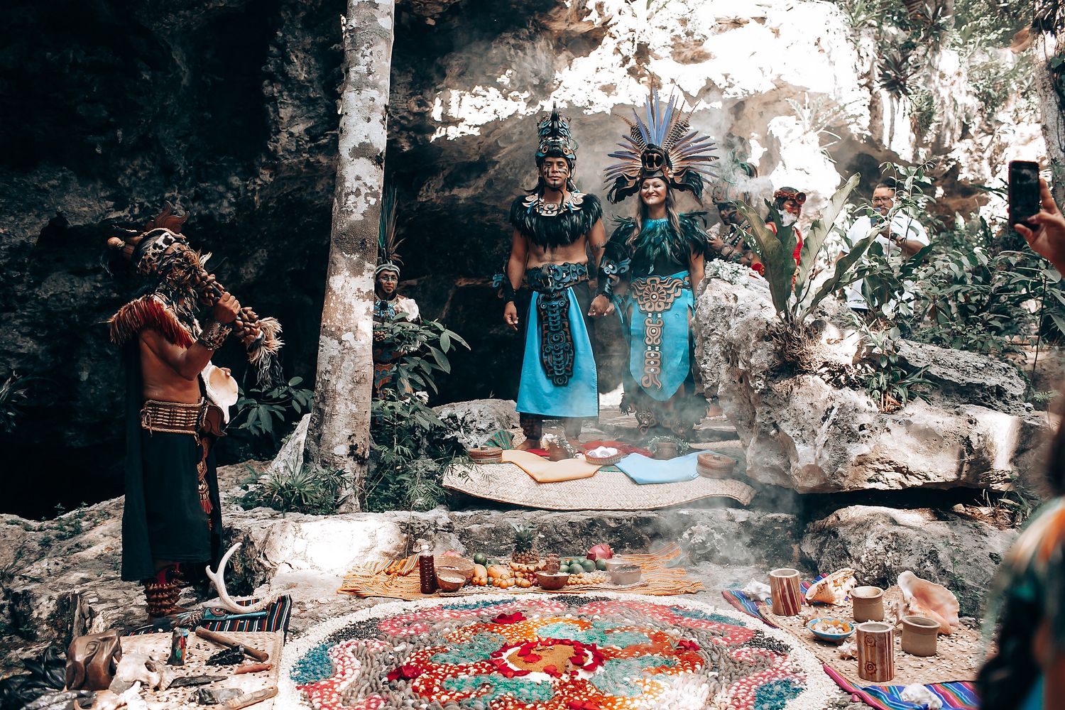 Elaborately costumed Mayan performers stand amid ritual offerings and colorful mandala on ground in ancient cave setting.