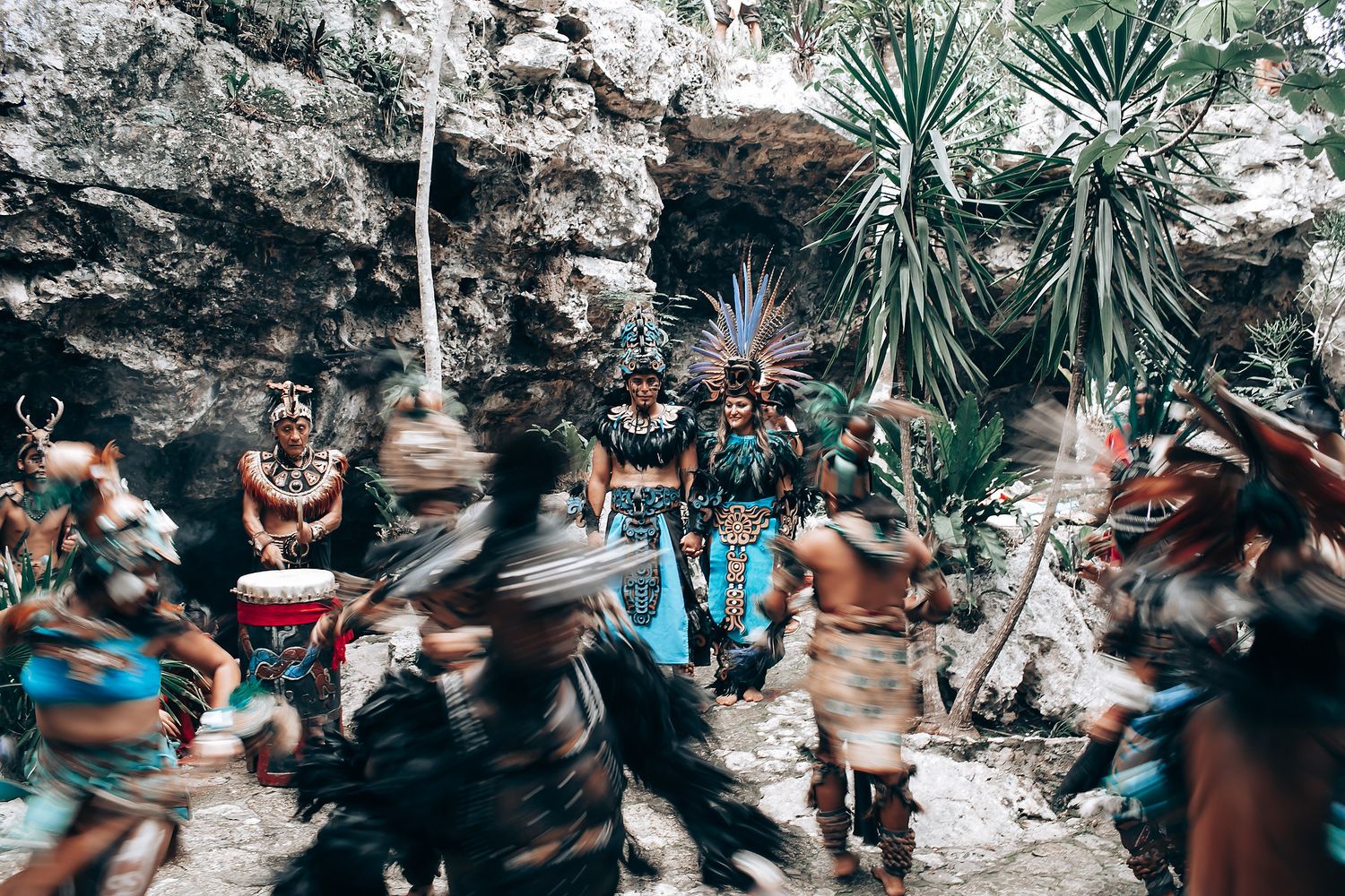 Dynamic group of Mayan ceremonial dancers in traditional costumes performing outdoors near rocky jungle terrain.