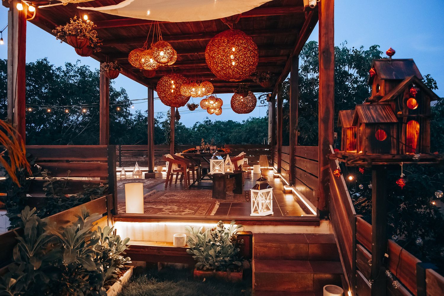 Cozy outdoor restaurant terrace at dusk with warm wicker pendant lights and string lights above dining tables.